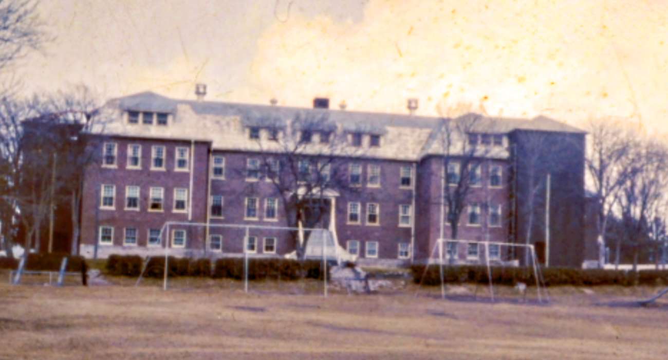 A photo of the exterior of Cecilia Jeffrey Indian Residential School, showing a red-brick building with a playing field and swings in front.