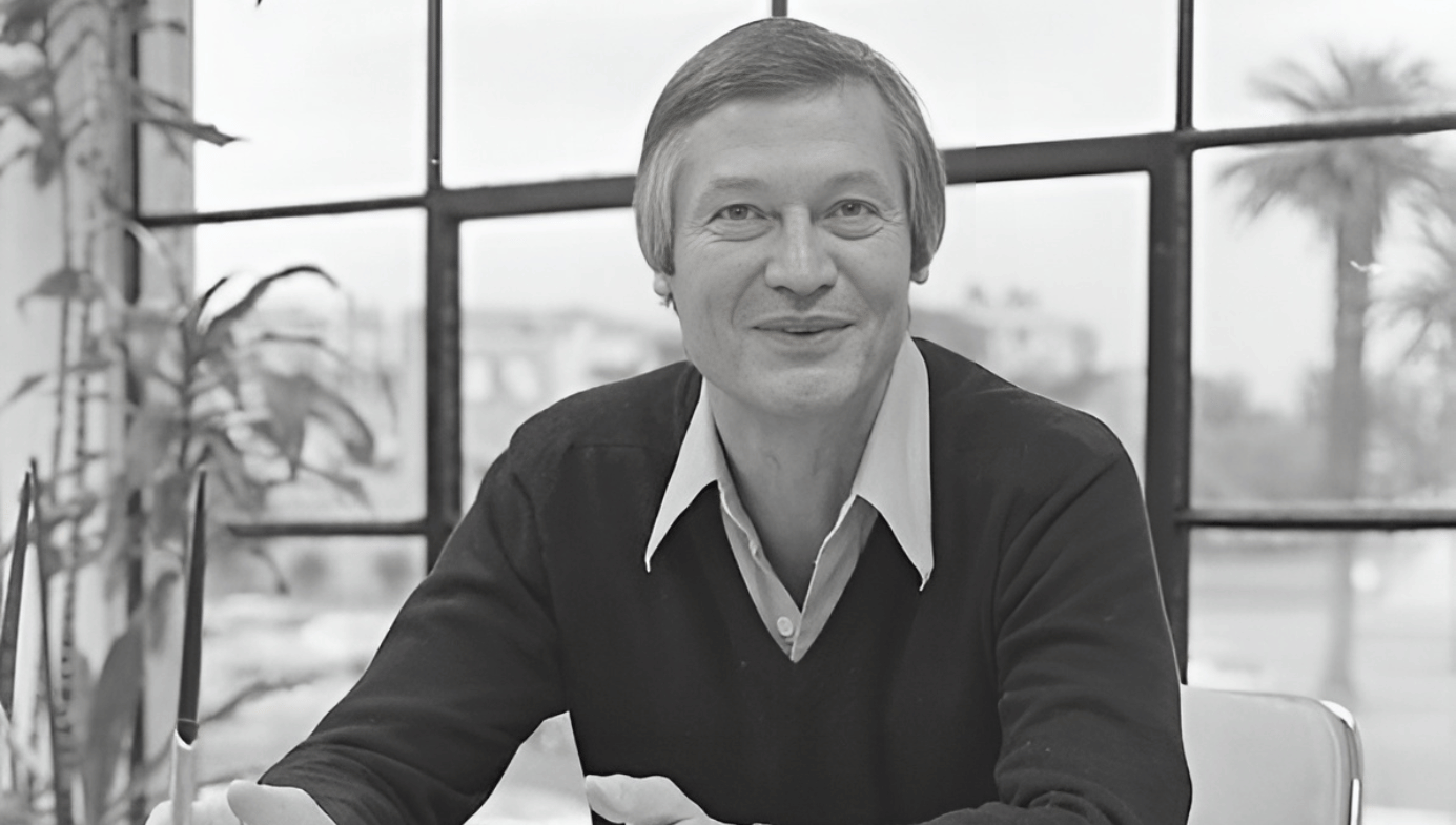 A black and white portrait photo of Roger Corman in 1978. He is sitting at a desk and looks happy. Palm trees in background. 