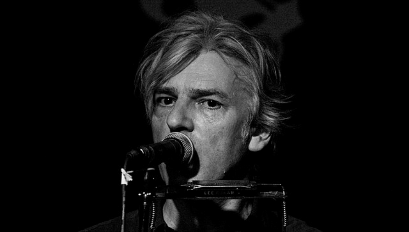 A close up of Robyn Hitchcock's face in black and white with a microphone covering his mouth.
