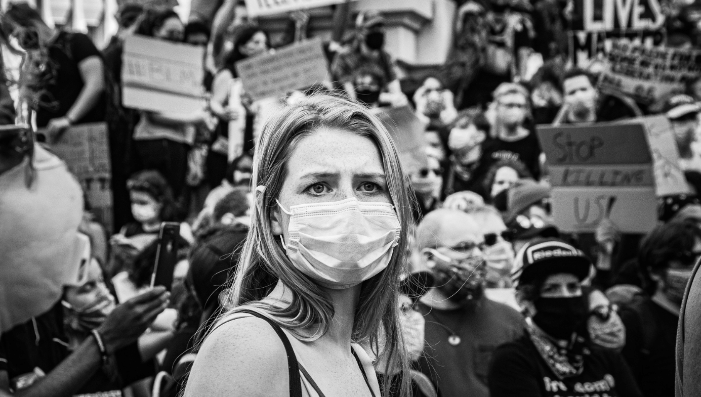 A young blonde woman with a COVID mask and worried eyes attends a BLM protester.