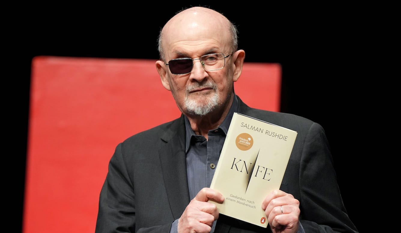 Salman Rushdie on stage, holding a copy of his book, Knife.