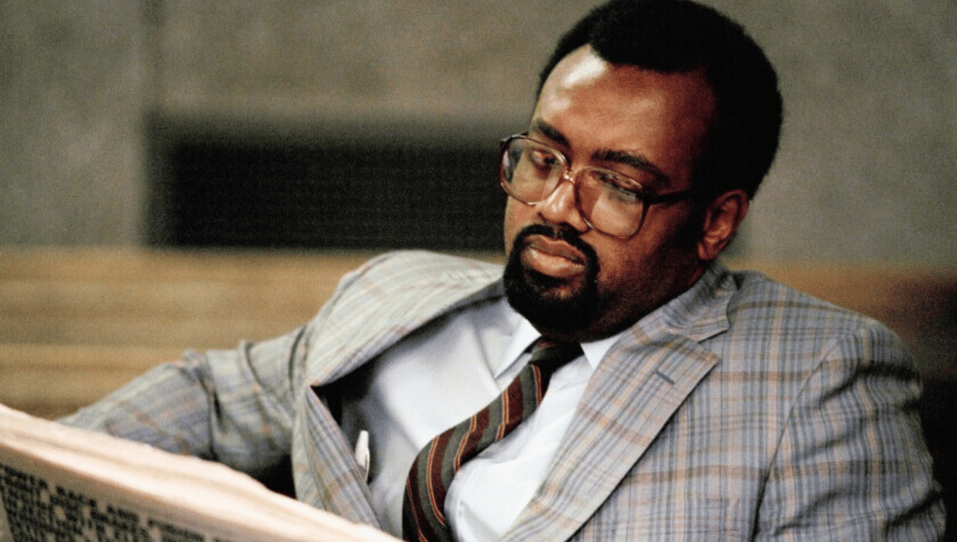 A photo of Glenn Loury reading a book in the 1980s. He is a black man with glasses, suit and tie