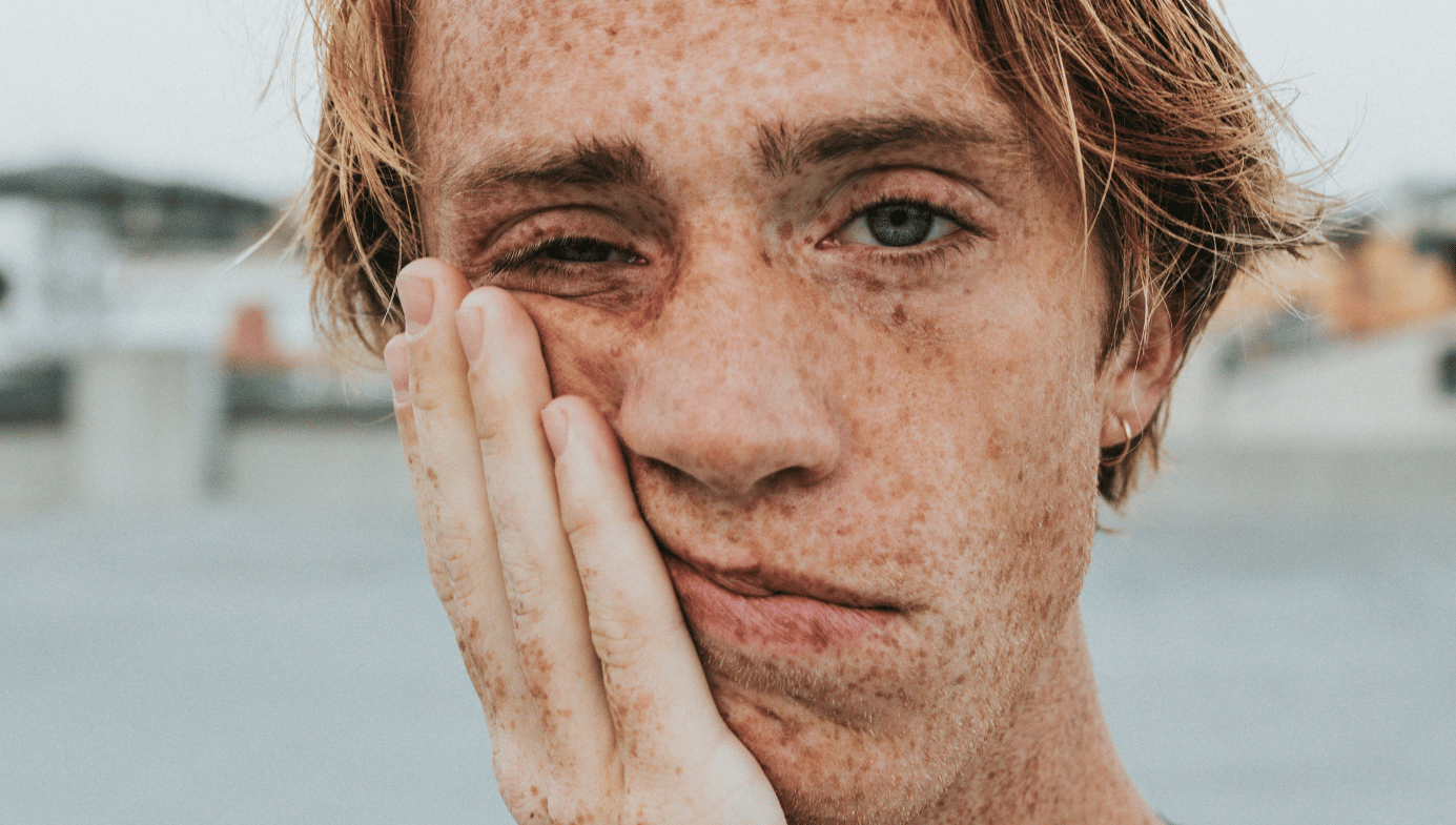 A freckly young man with blue eyes and blonde hair holds his hand to his face and looks sad.