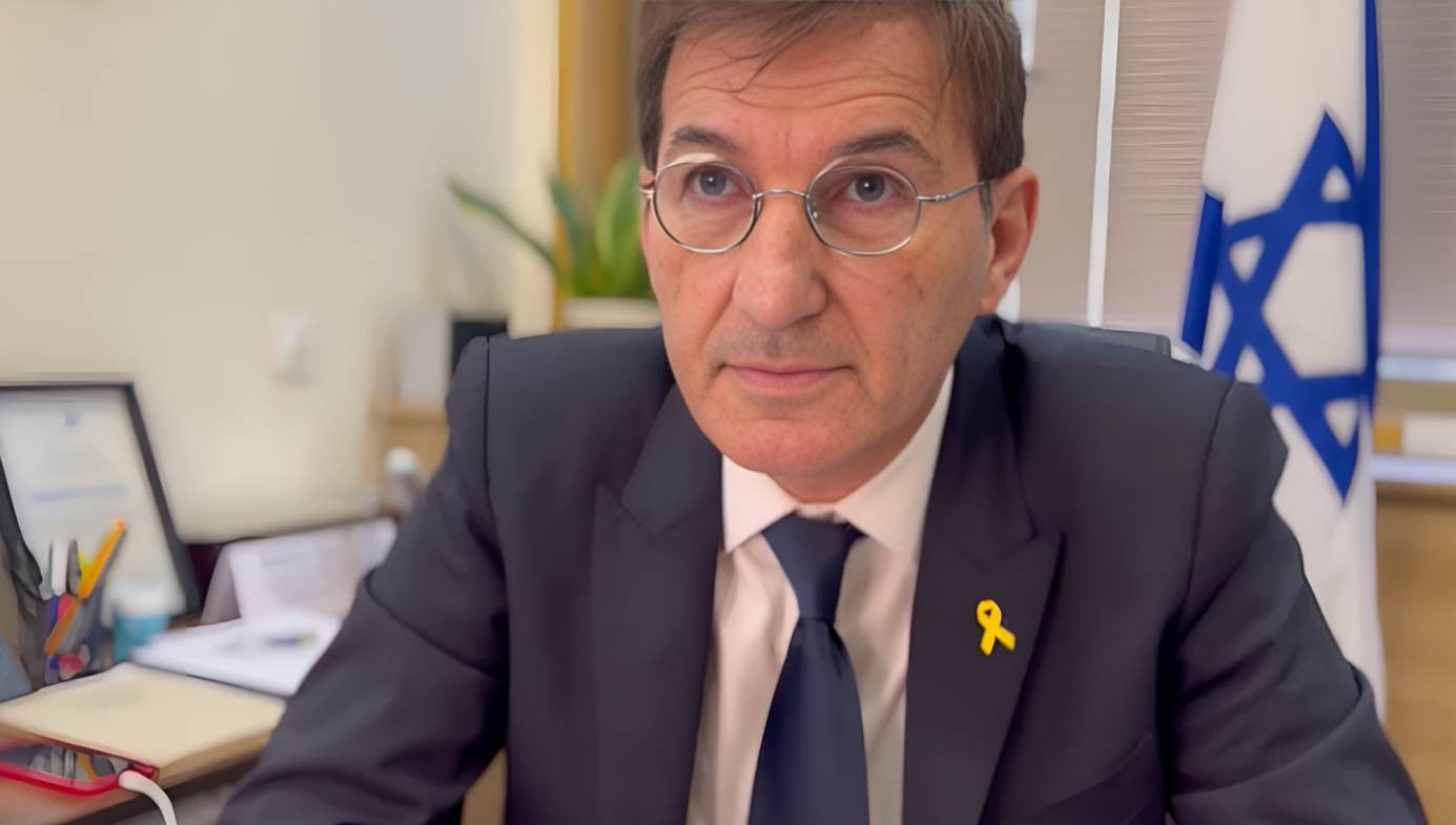 Boaz wears round glasses, a suit and tie, and a yellow lapel ribbon. An Israeli flag hangs in the background