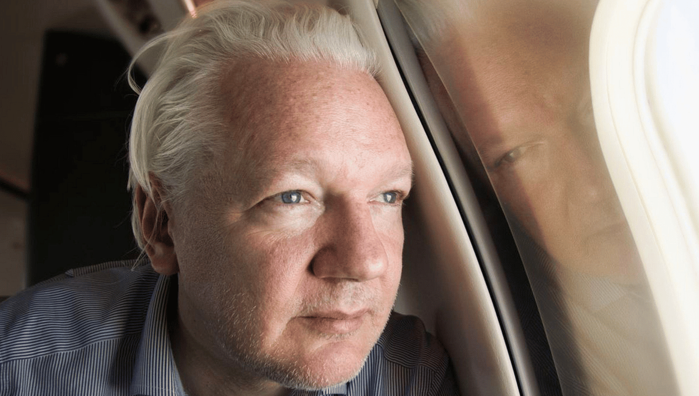 Julian Assange looks out the window of an aeroplane. You can see his reflection mirrored in the window. 