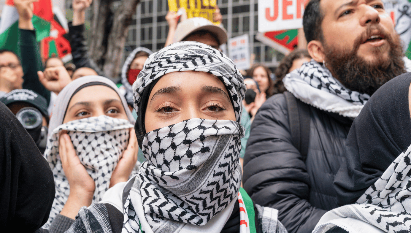 A young woman in a keffiyeh wrapped around her face. She is in a crowd of pro-Palestine supporters