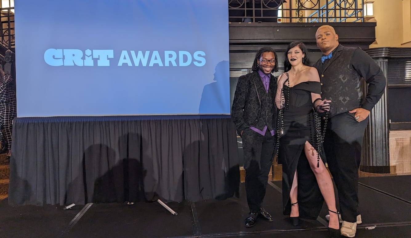 A woman and two men on stage in front of a poster at the GRIT awards. 