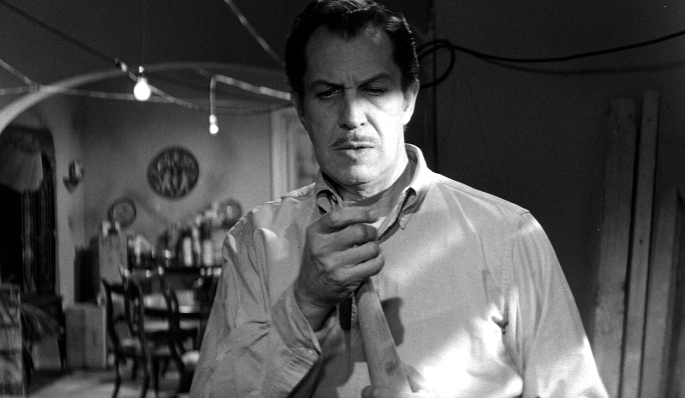 Vincent Price in a black and white film still, holding a wooden object, with a dining table in the background