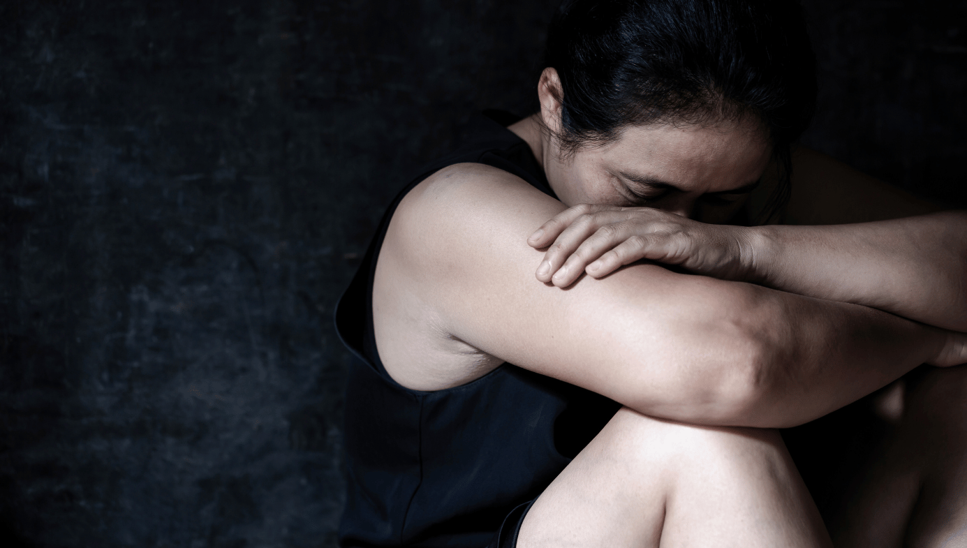 A woman looks sad as she sits on the ground against the wall and hugs her body.