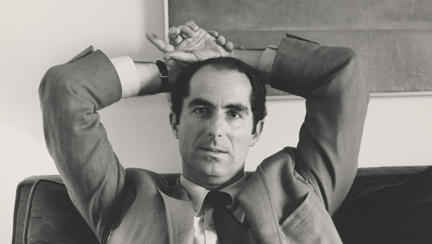 Philip Roth, seated, hands behind head. He's in a suit. Dark hair and thick eyebrows.