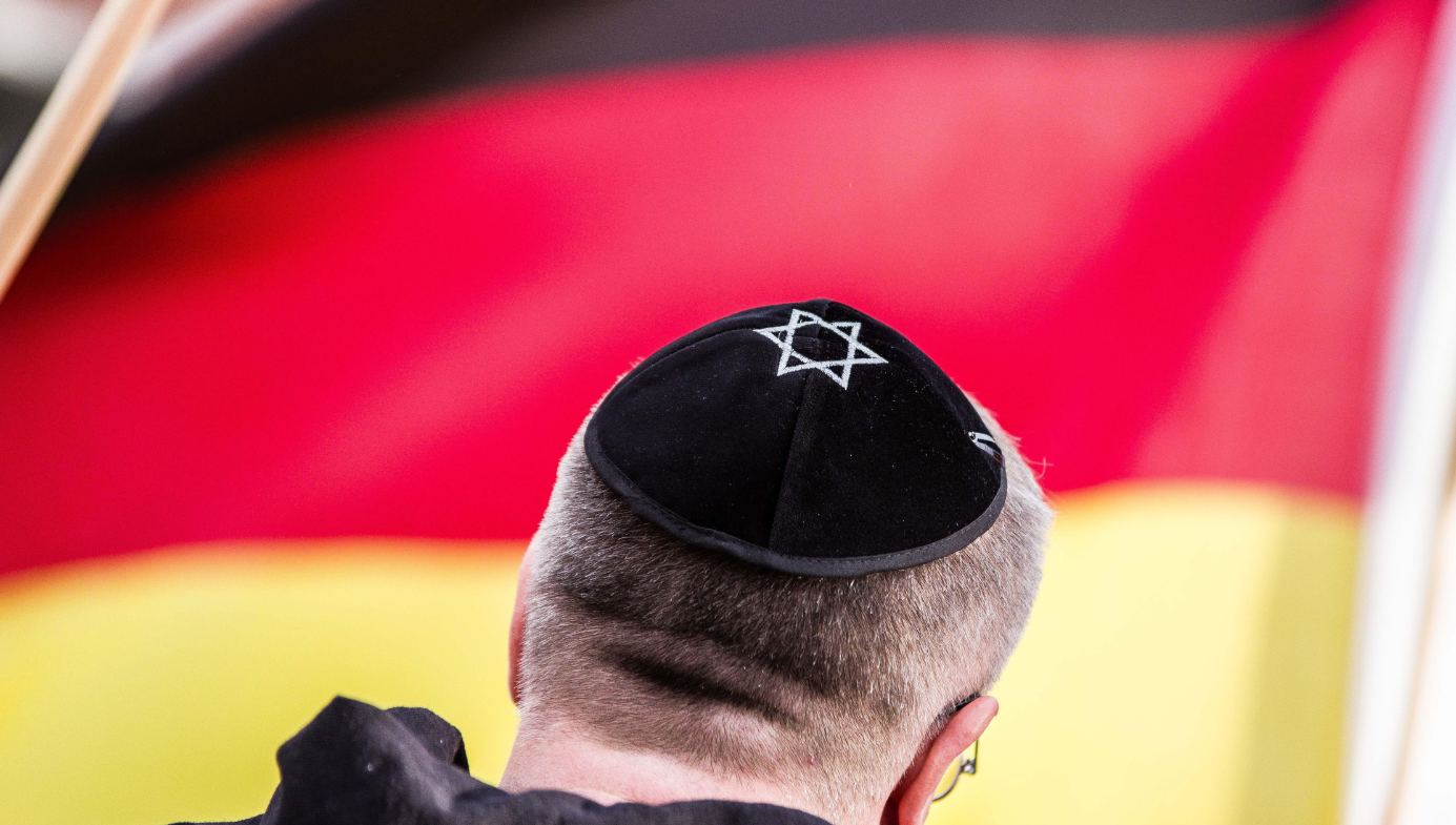 The back of a middle-aged white man wearing a black yarmulke with a star of David embroidered on it. Backdrop of German flag.