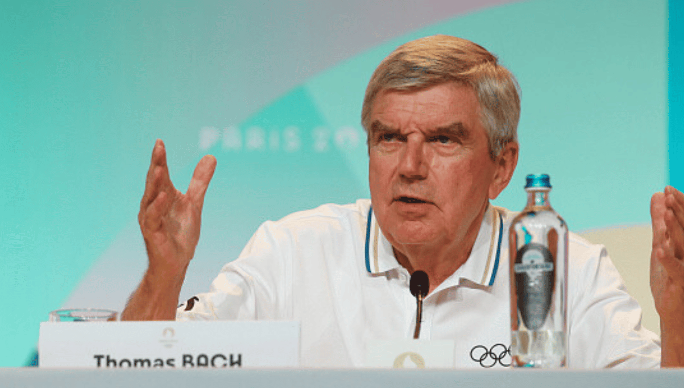 Thomas Bach, President of the International Olympic Committee, at a podium with a water bottle, speaking.