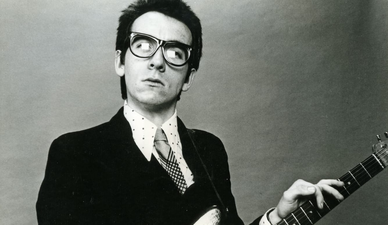 Elvis Costello, black and white photograph, in glasses, a shirt, jacket and tie, fingers on the neck of a guitar. 