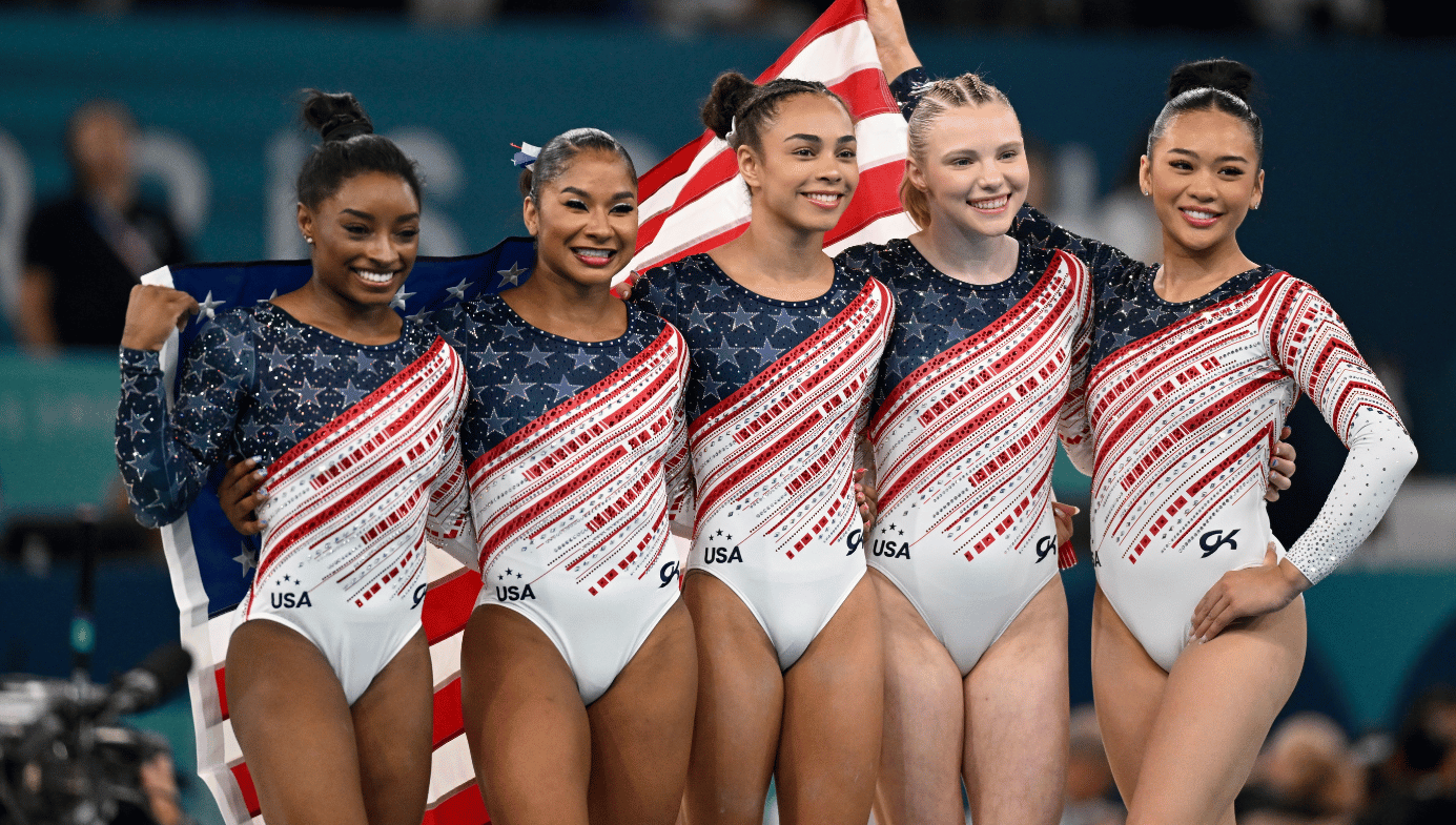 US Olympics Women's Gymnastics Team in their costumes, in front of an American flag. 