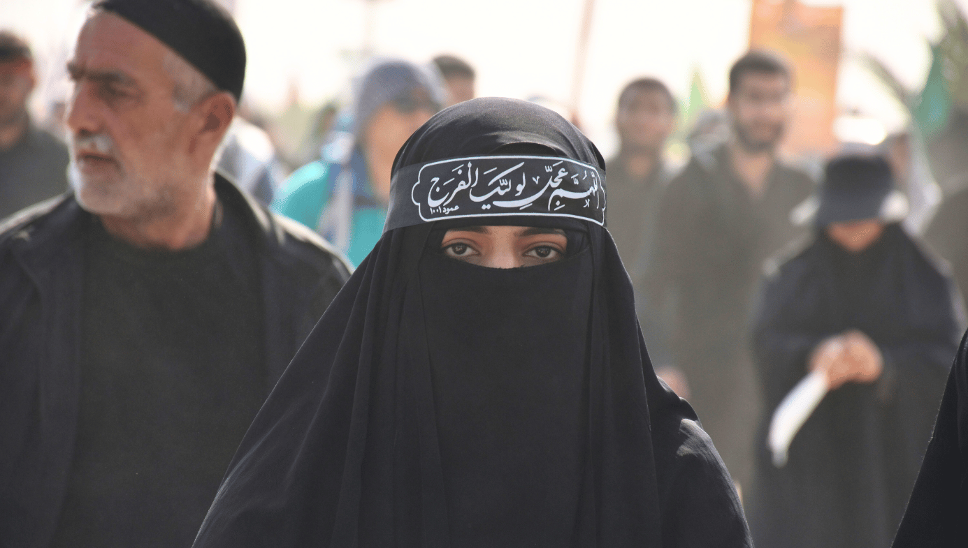 A woman in a burqa with Arabic lettering on her headband looks at the camera. Men in the background.