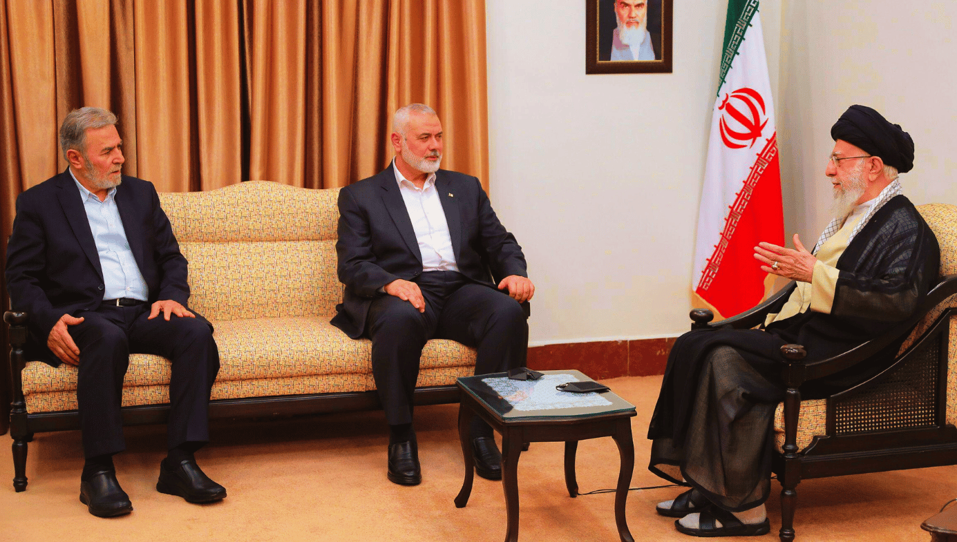 Two men in suits sit on a couch facing an ayatollah in robes on a chair. There is an Iranian flag next to him. 