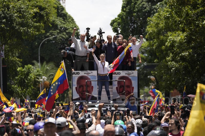 Maria Corina Machado and allies standing on a truck above a crowd of supporters. She is holding a Venezuelan flag. 