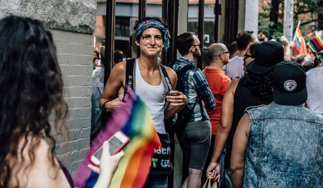 Lauren Lubin at a protest, smiling at the camera, in a tank top, with a bandana, waving a rainbow flag. 