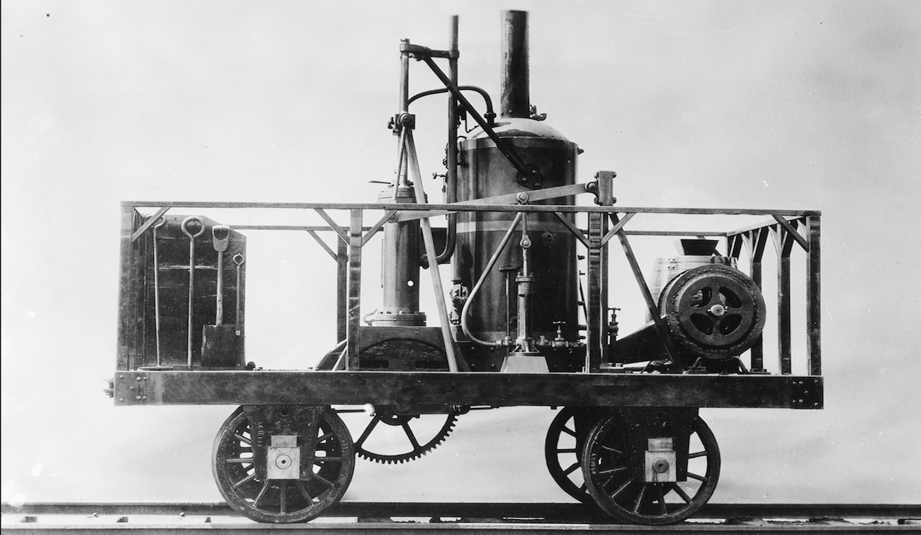 Black and white photo of a replica of an early steam-powered locomotive, on train tracks, against a blank wall. 