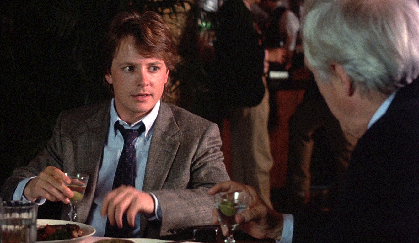 Michael J. Fox in Bright Lights, Big City, in a suit, at a restaurant, talking to an older man, holding wine.