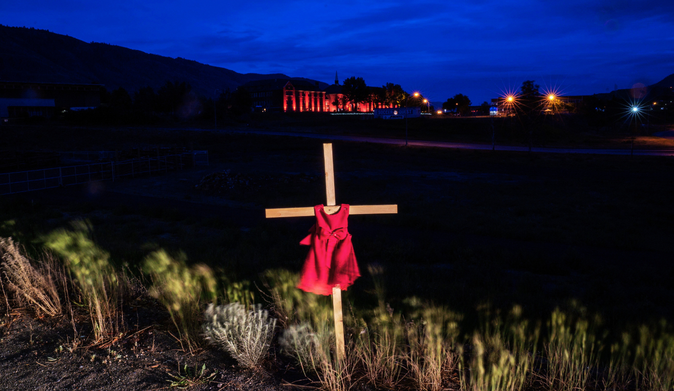 Red child's dress on a wooden cross at night. Kamloops school lit up in red in the background. 