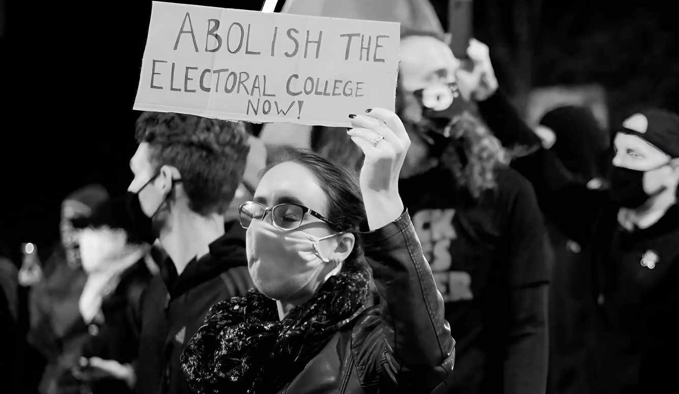 A woman in a mask and glasses holds a sign saying ABOLISH THE ELECTORAL COLLEGE NOW! at a rally.