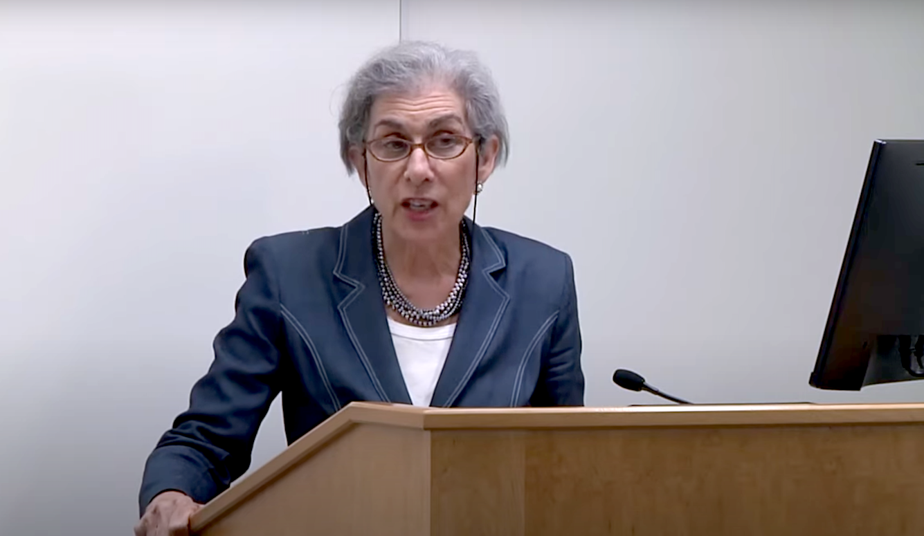 Amy Wax at a podium. She is a grey-haired lady with glasses in a dark blue jacket and white T-shirt. 