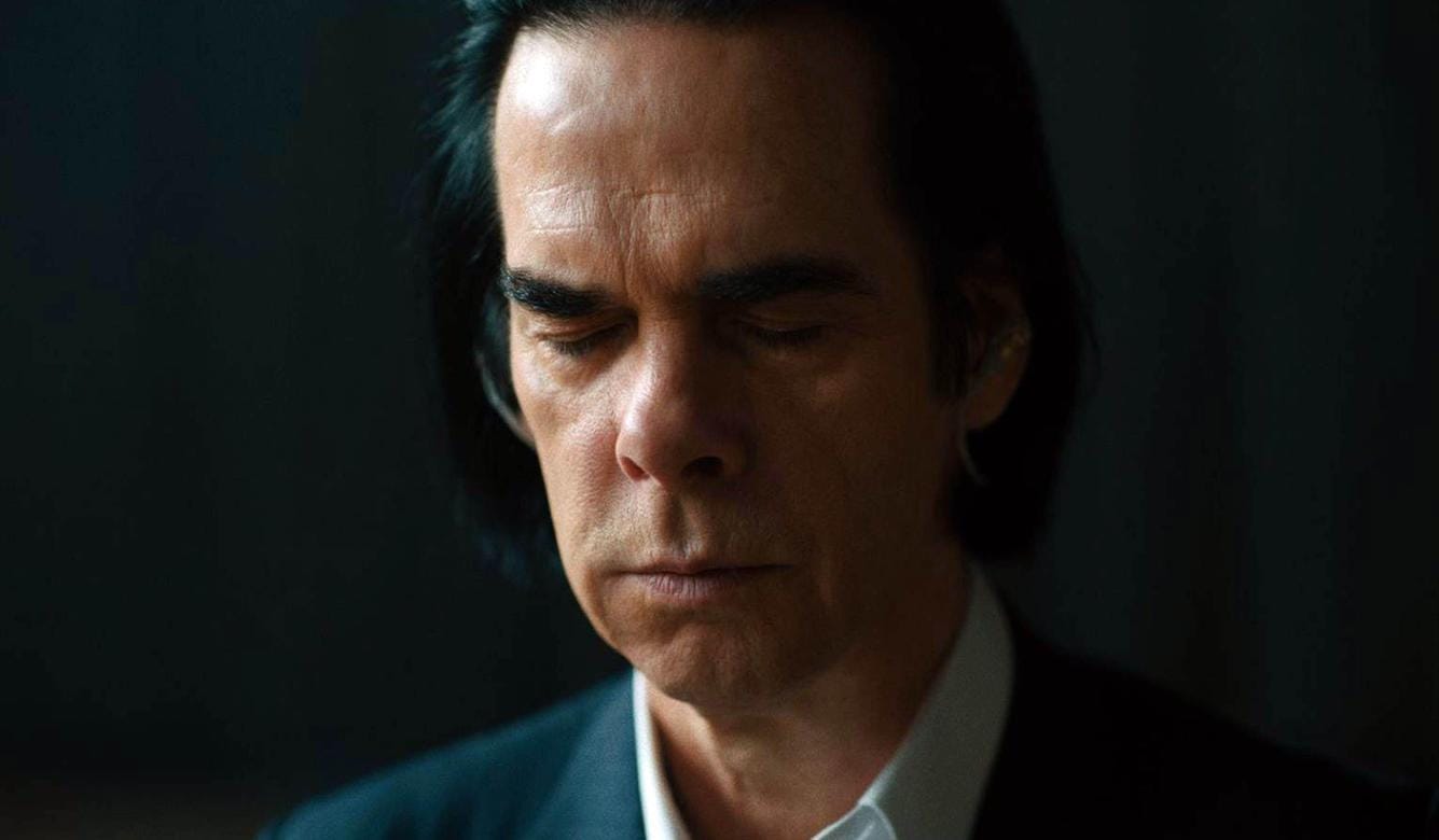 Nick Cave, a striking man in his 50s with black hair , eyes closed, wearing a blue jacket and light blue shirt. 