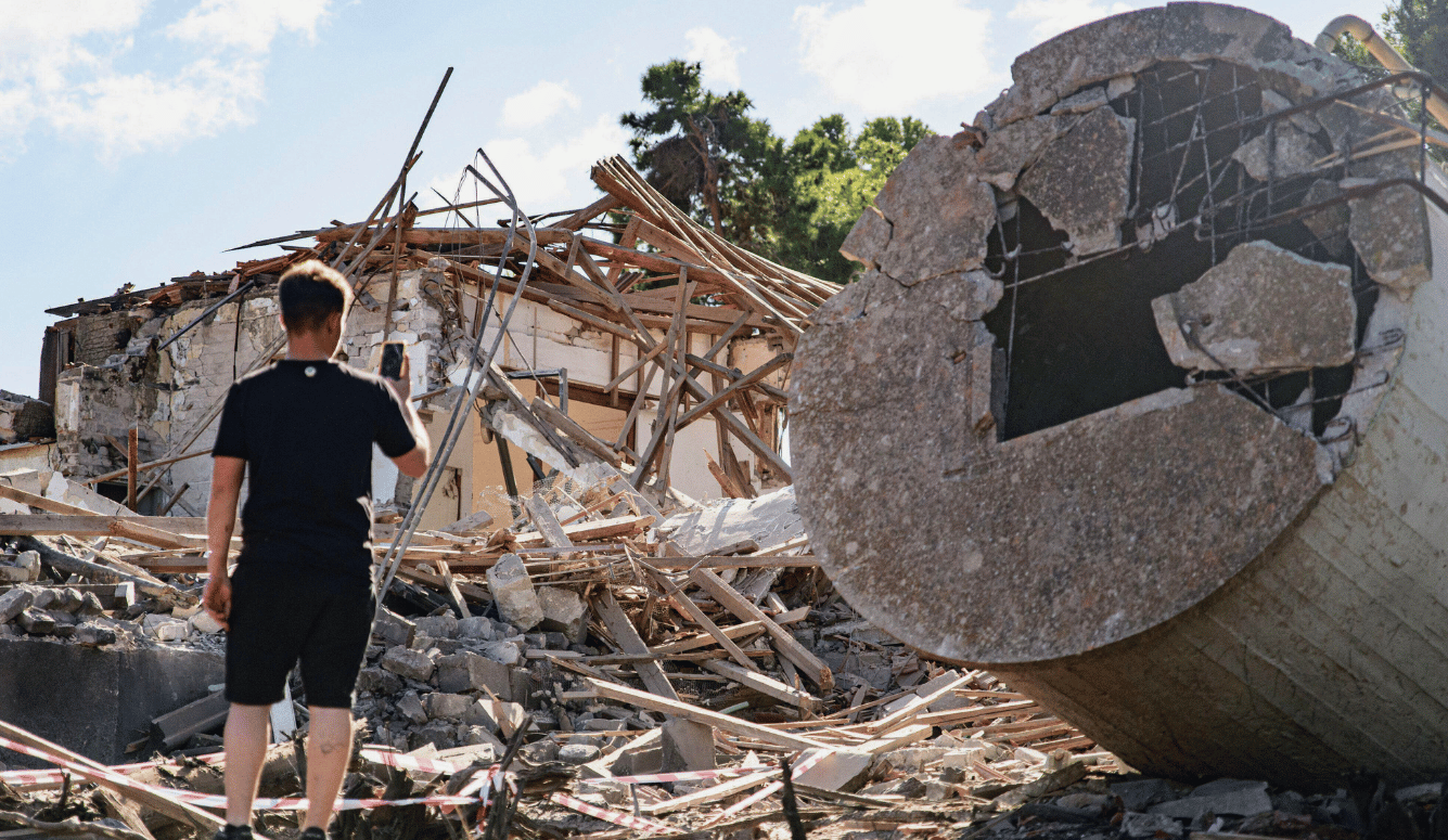 A destroyed building and half a missile. A man takes a photo of the wreckage on his phone.