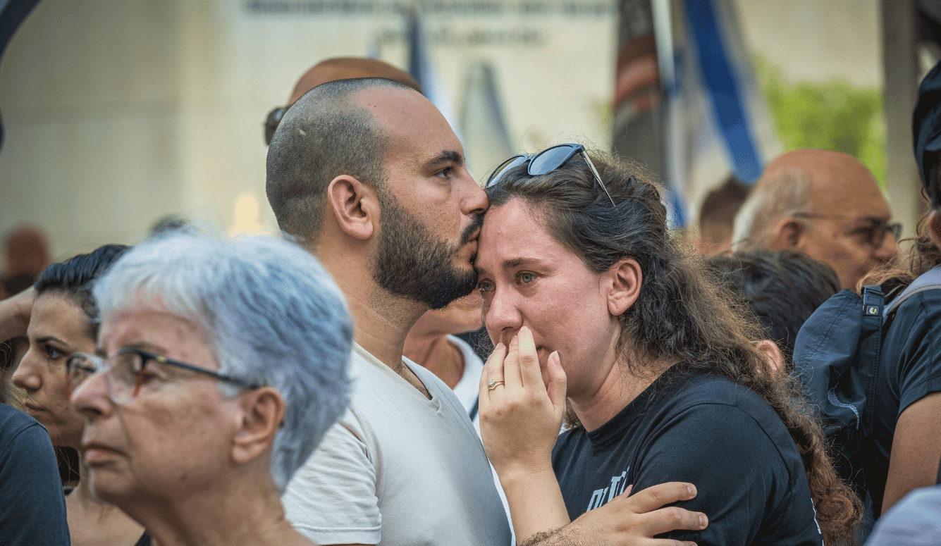 A crowd of people, mourning. Man comforts a crying woman in the centre. 