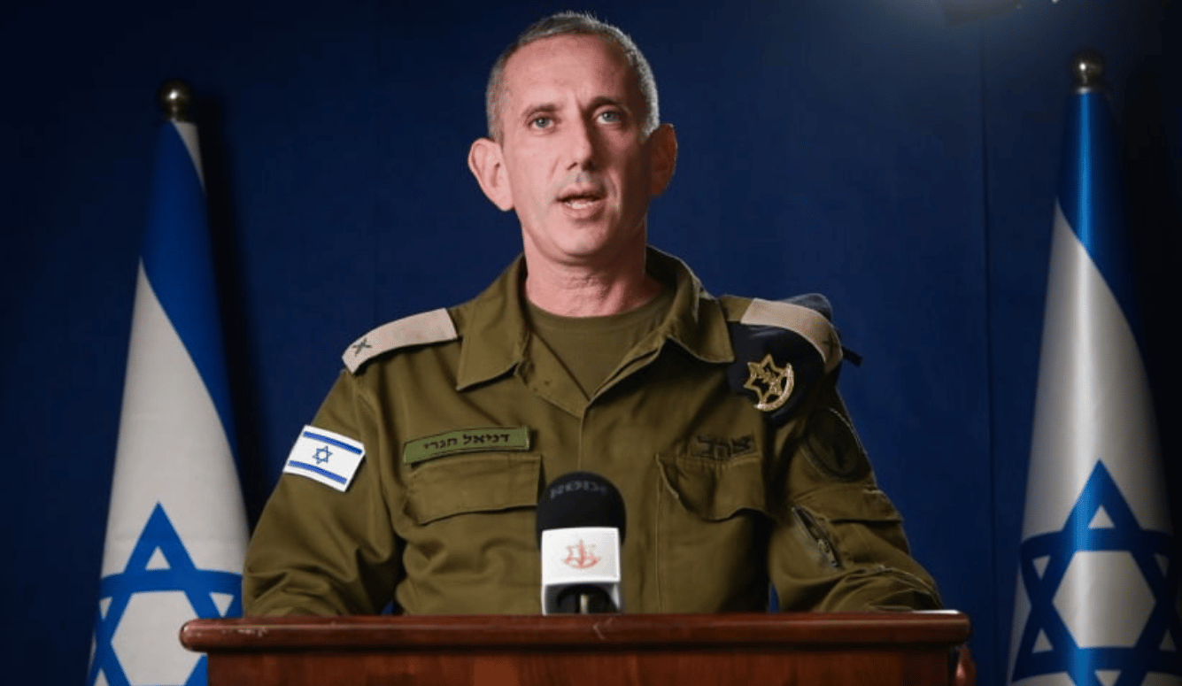 Daniel Hagari, a middle-aged white man with blue eyes in a military uniform speaks at a podium with Israel flags behind him.