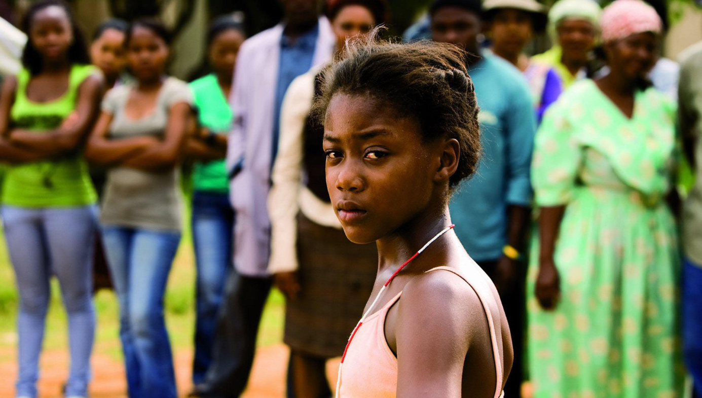 A young African girl looks at the camera, defiantly. A group of people stand behind her, at a slight distance. 