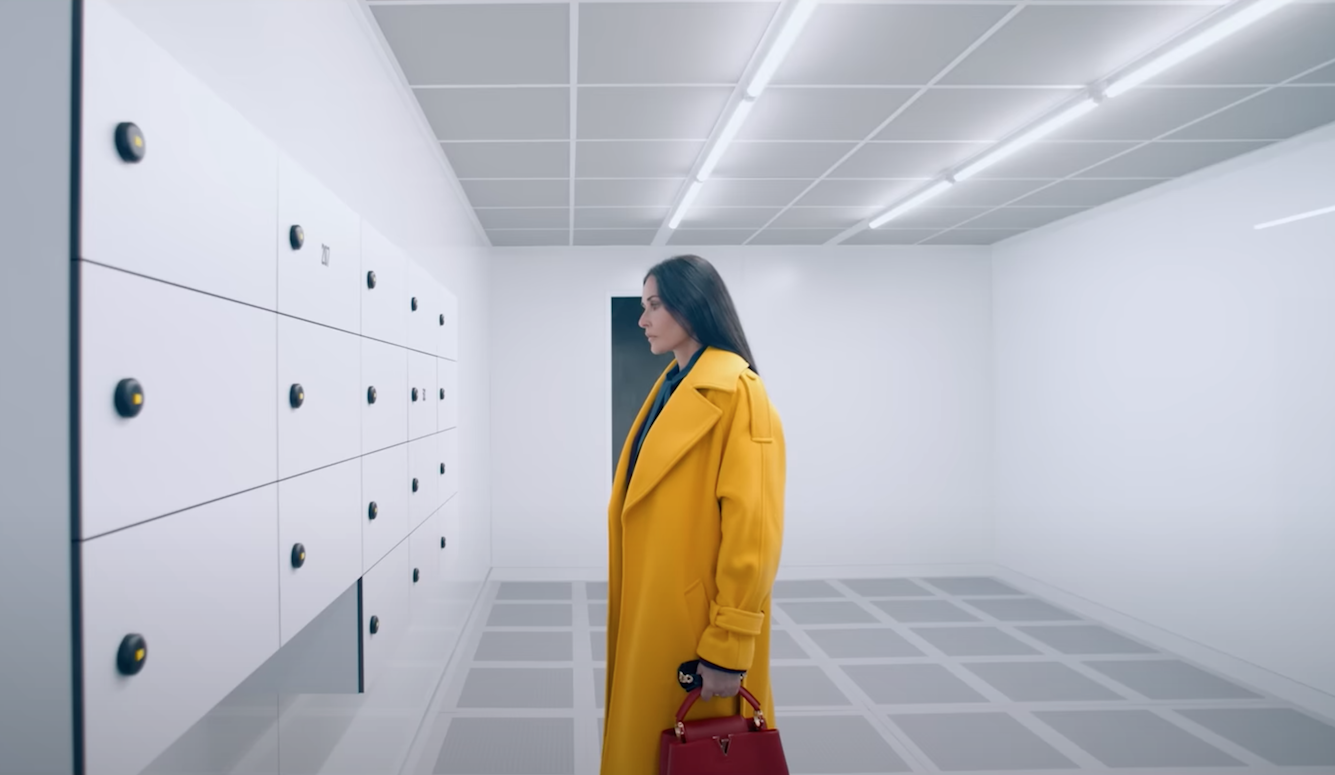 A woman in a yellow coat stands in front of a row of lockers. She is attractive, fifties, carries an expensive handbag. 