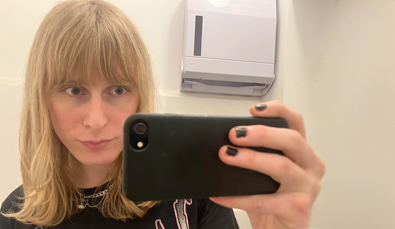 The face of a trans woman, dressed in a feminine manner, looking into a mobile phone, in a public toilet. 