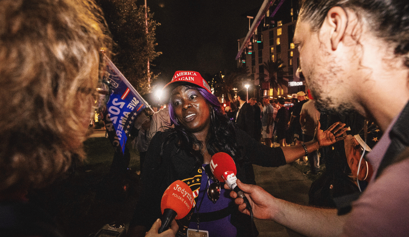 Black female Trump supporter wearing MAGA merch speaks into two microphones. 