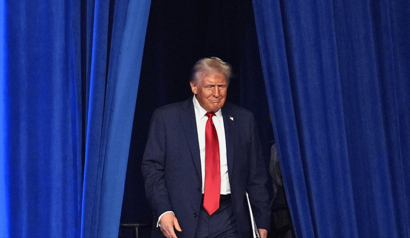 Trump in a red tie walks through blue curtains looking chuffed.