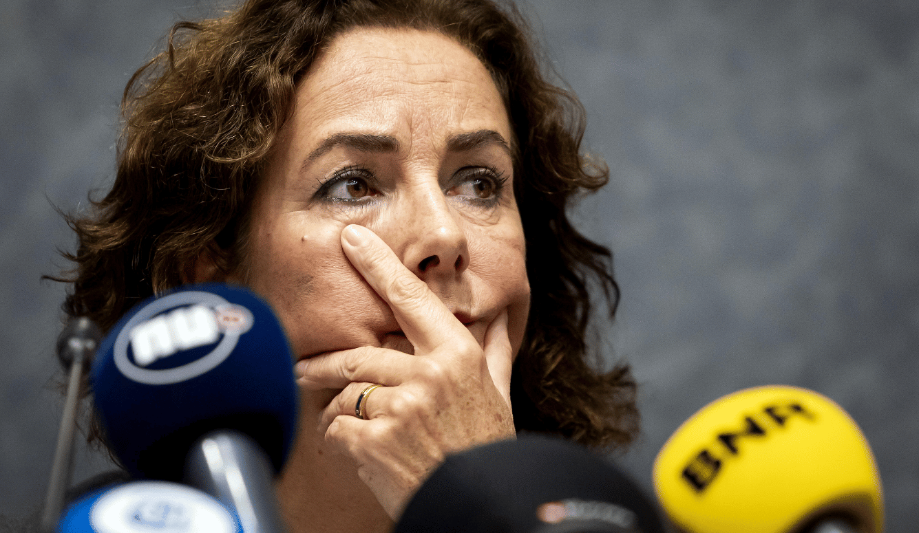 A brunette middle-aged female mayor of Amsterdam looks concerned with her hand over her mouth and microphones pointi