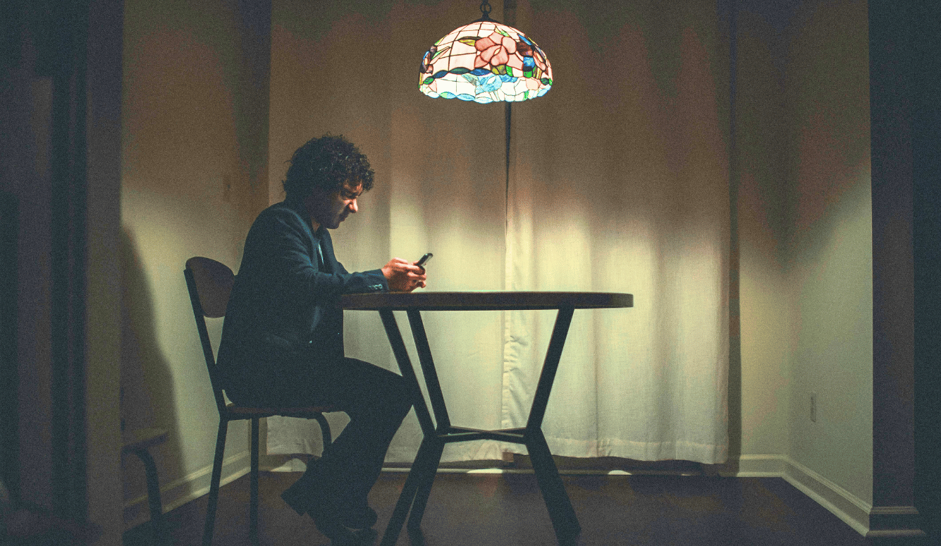 Man in black shirt sitting on a chair alone in lamp light. He's on his phone. He looks lonely.