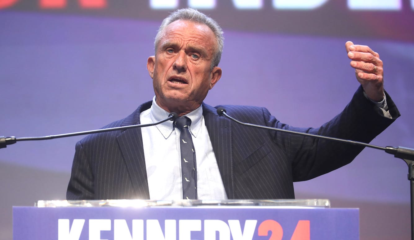 RFK Jr. is an elderly white man with ruddy skin and blue eyes. He is speaking at a podium, wearing a suit.