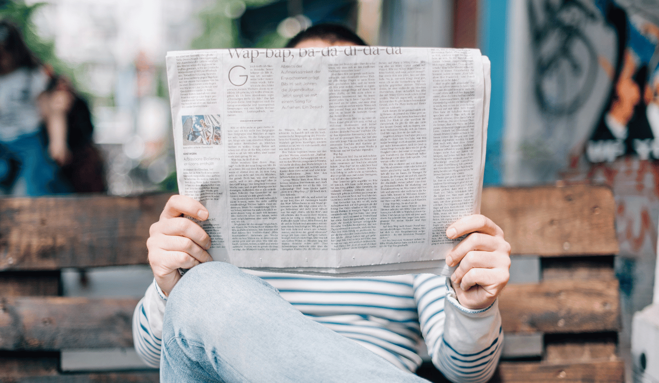 A man in a Breton T-shirt and jeans sits on a bench reading a newspaper.