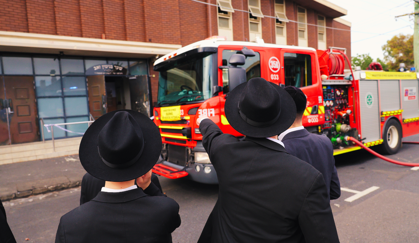 Jewish men in black hats and jackets watch a fire engine pull up to a synagogue.