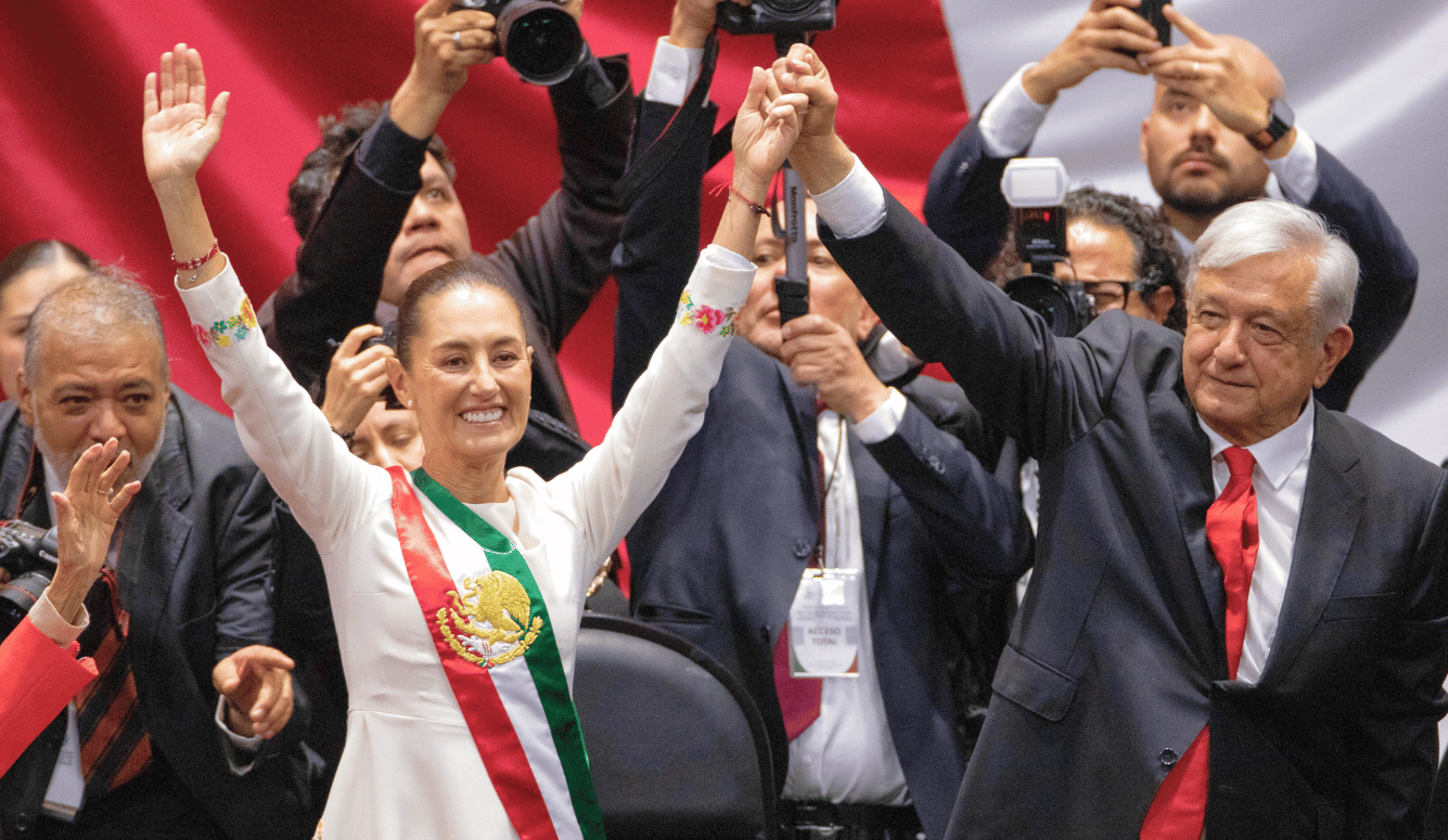 Claudia Sheinbaum Pardo, wearing a white dress, holds raised hands with Andrés Manuel López Obrador. Paparazzi in the backg 