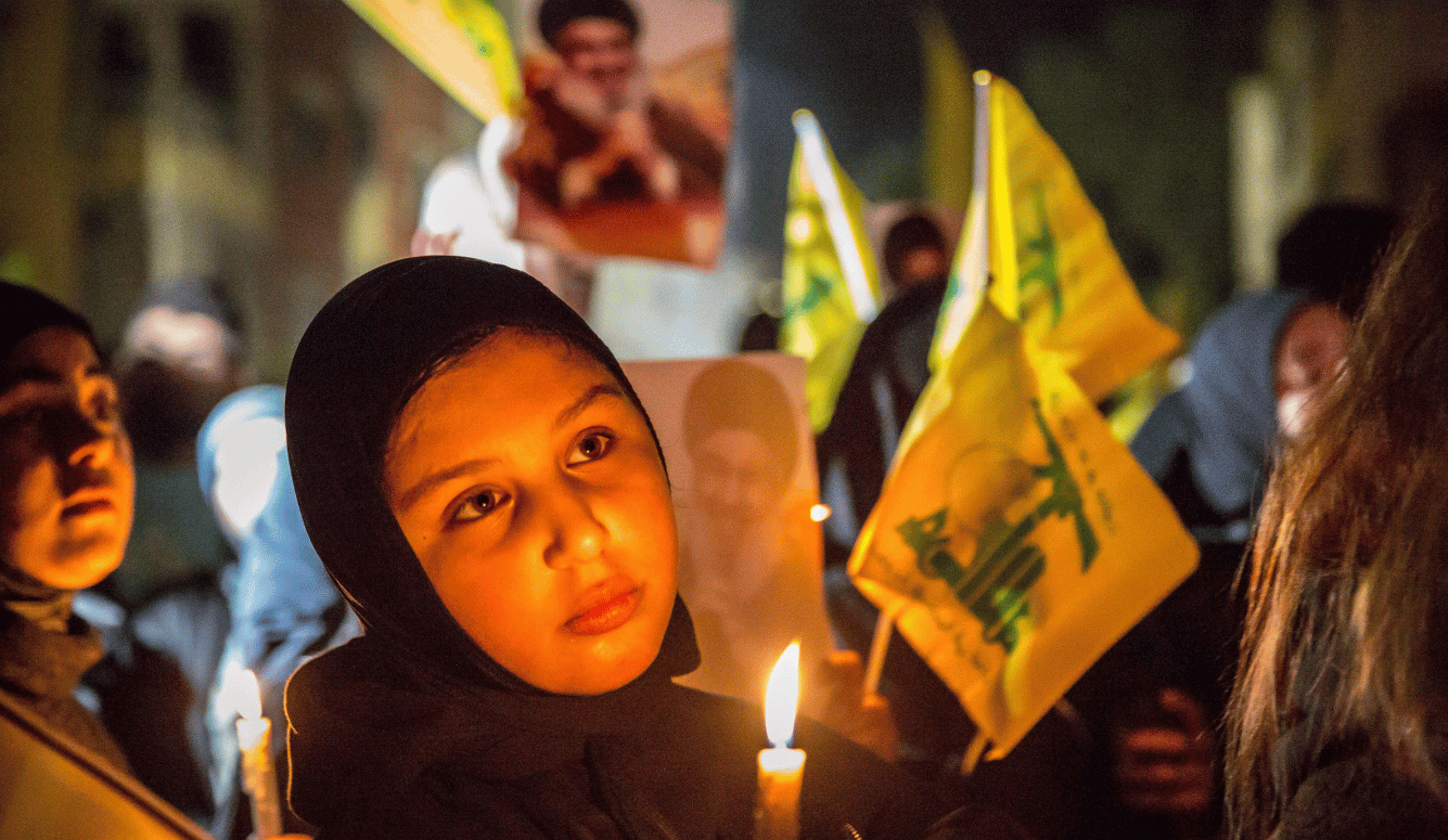 A young woman in a hijab holds a candle and Hezbollah flag. She looks sad.