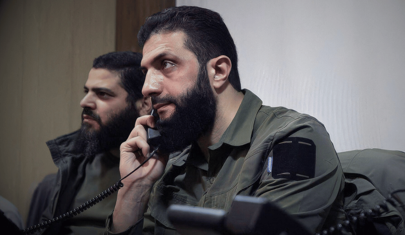 Ahmed Al-Shara is a bearded pale-skinned man. He is speaking into a telephone, wearing army fatigues. 