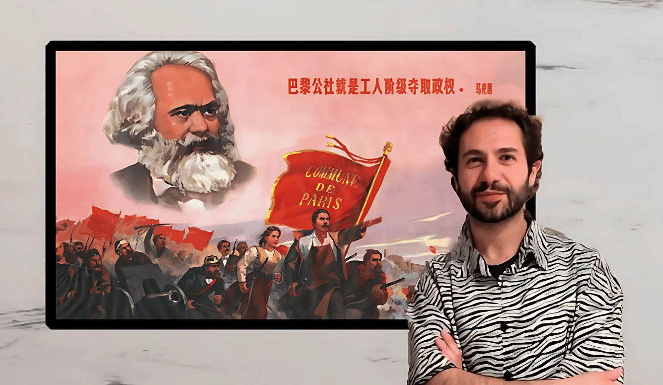 A dark haired young man with a beard smiles in front of a painting of Marxc