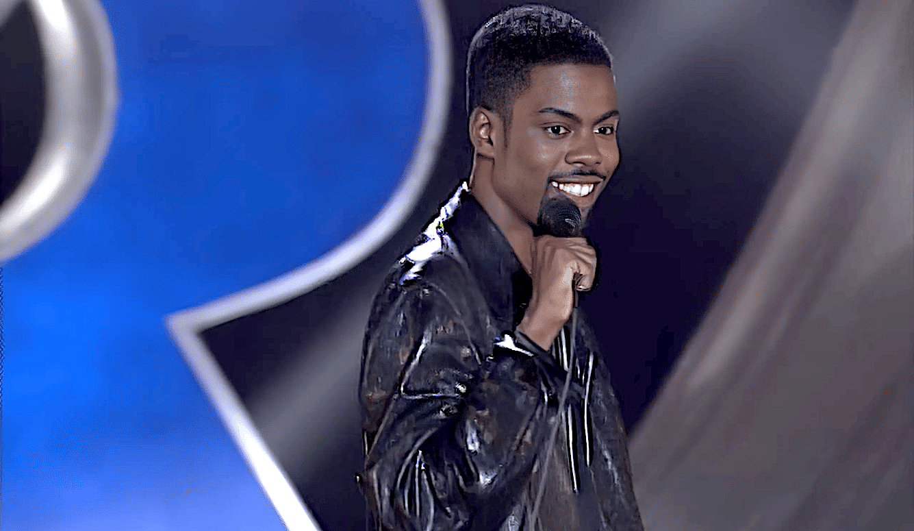 Chris Rock at a microphone. He is a young black man, smiling, wearing a shiny black shirt. 