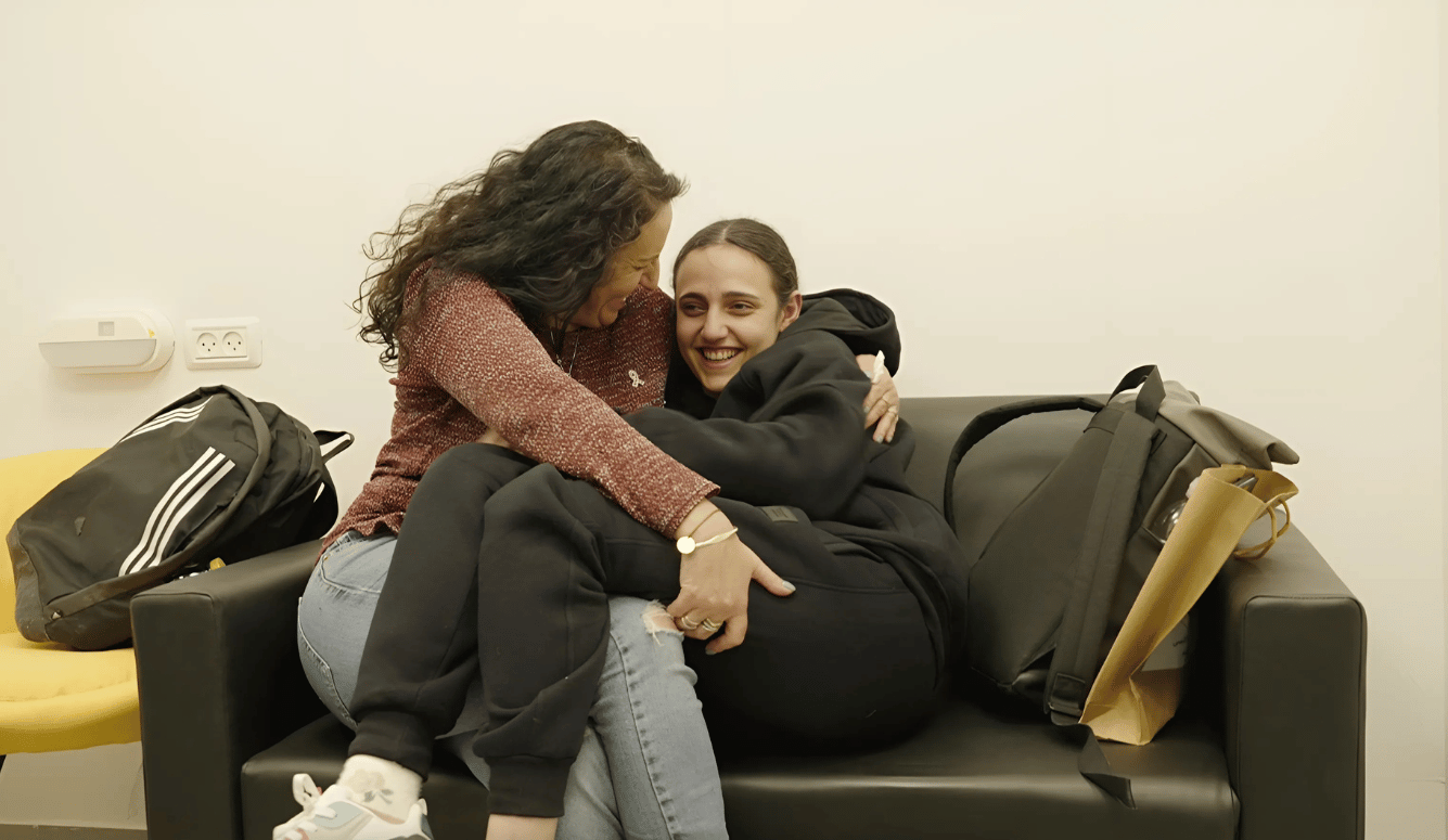 A young woman in a tracksuit is embraced by her mother, in jeans, on a couch. 