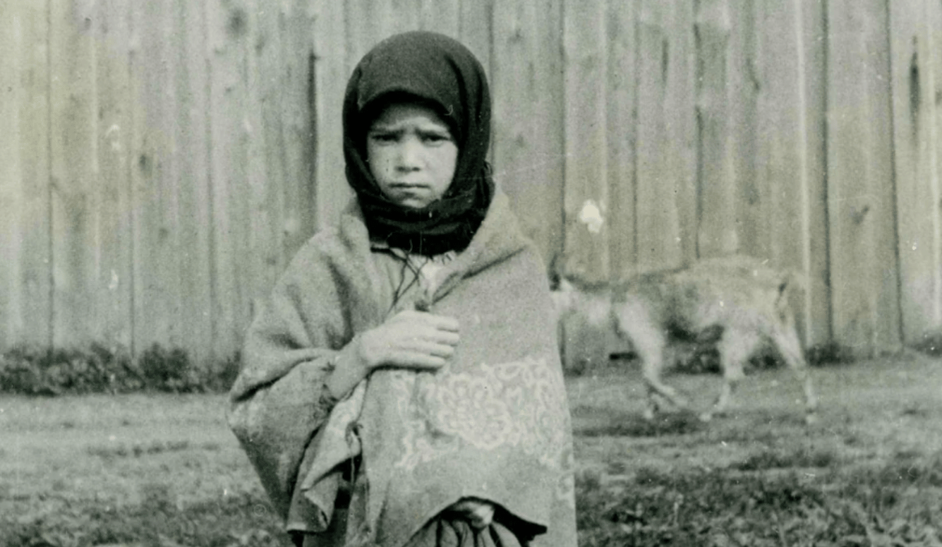 A young girl in a headscarf and shawl looks sad and starving.