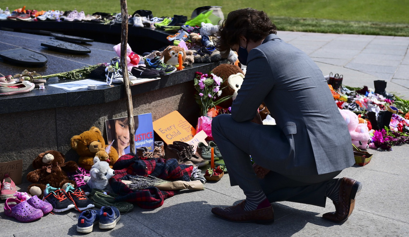 Justin Trudeau kneels in front of a memorial with flowers, teddy bears and children's shoes. 