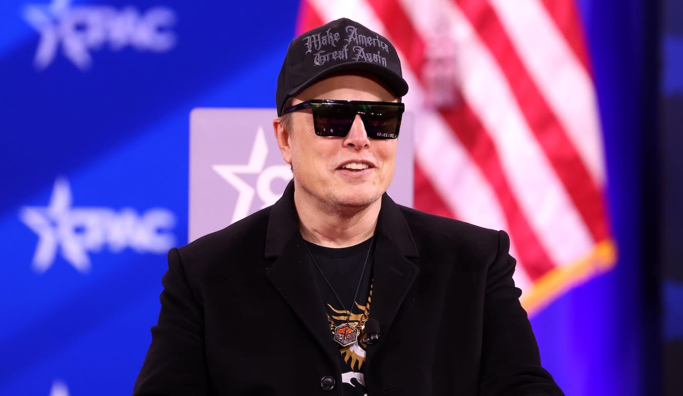 Elon Musk in sunglasses in front of an American flag and the CPAC logo